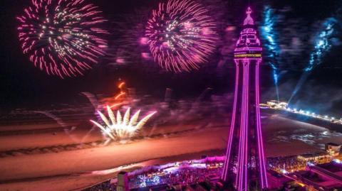 Blackpool Tower and the beach lit up in pinks and purples as fireworks blast against the night sky