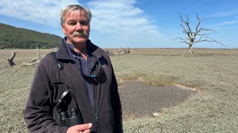Nigel Hester is standing in the middle of the salt marsh wearing a black fleece. The are is quite bare with some grass and a bare tree. 