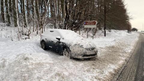 A car covered in snow at the side of a rural road.