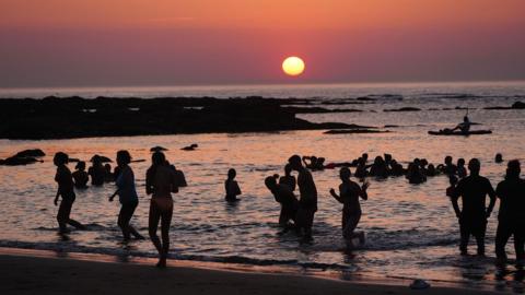 People in the sea silhouetted against a purple and orange sky 
