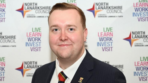 A man in a suit standing in front of a North Lanarkshire Council backdrop featuring repeated text promoting live, learn, work, invest, and visit