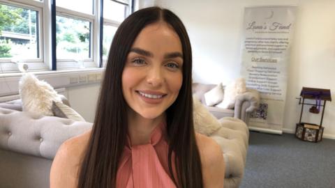 Aimee Conroy pictured in a building with white walls with windows over her right shoulder and a pair of light grey sofas behind her. Ms Conroy is seen wearing a pink sleeveless top with long dark hair.