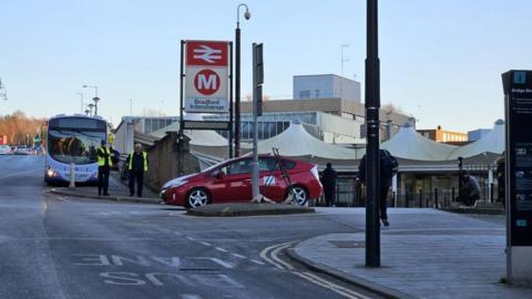 A red car drives out of Bradford Interchange, with two uniformed personnel standing beside a bus on the road. The interchange building with white canopy structures is visible in the background, along with signage for rail and metro services. A few pedestrians are walking nearby.