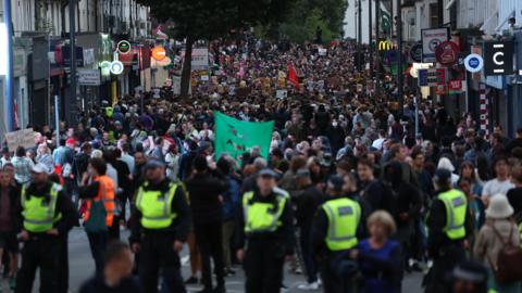 A large crowd of protestors filling a High Street with a line of police in high viz jackets in the foreground