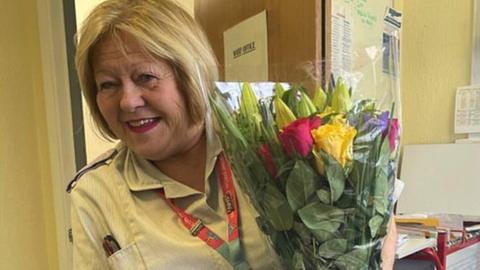 Image of Sue Sheriton holding a bunch of flowers. A card reading Cheers to 45 Years! is in front of her, along with a chocolate cake.