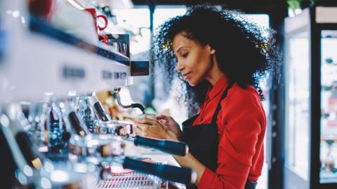 Young female barista wearing a red shirt and a black apron using espresso maker at café.