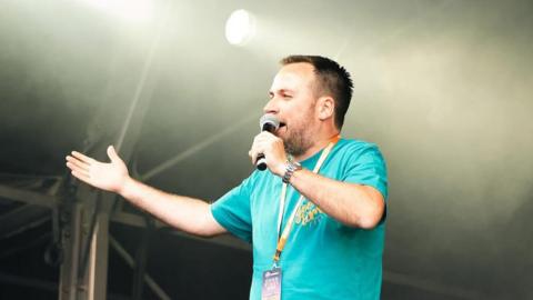 Man wearing blue top with yellow lanyard stood with a microphone on stage.