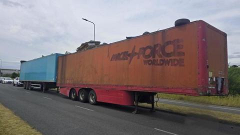 Two trailers - one blue and one orange with a faded Parcelforce Worldwide logo on - parked on a road.