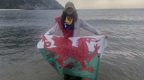 Man with long hair is wearing a cream bucket hat and standing in the sea up to his knees. He's weaing a cream long-sleeved shirt and holding the Wales flag in front of him. He also has the flag of Venezuela around his neck.