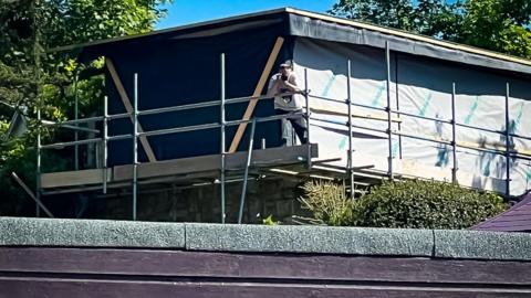 the tower looms above a garden wall. It is above a roof in the right corner of the picture and is surrounded by scaffolding, with black plastic sheeting on the walls. A man in a baseball cap, with white t shirt and dark trousers is leaning on the scaffold rail looking towards the neighbour's property. He has a phone held up near his face with both hands.  