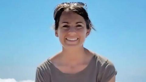 Family handout image of Lisa Ross - Close-up portrait of a person outdoors against a clear blue sky, wearing a grey short-sleeved top with sunglasses resting on the top of the head. The image is brightly lit by sunlight, with a small section of white cloud visible in the lower left corner.