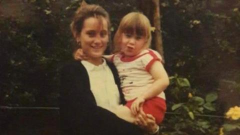 Natalie Pearman holding her younger sister Georgina Simpson in her arms. Natalie as tied up brunette hair and is wearing a dark jumper and white shirt. Georgina is much younger, and is wearing a white and red outfit.