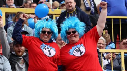 Two women are dressed in costumes similar to Thing One and Thing Two characters from Dr Seuss's Cat in the Hat books. They wear blue wigs with red tops and sunglasses One of the women's tops says Amy 1 and the other says Amy 2. They are standing in football stadium stands with their arms raised in celebration as they smile at the camera.