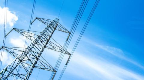 A view up towards a steel electricity tower. The tower is tall structure made from metal beams. Thick cables are suspended from "arms" on the tower. The pylon is pictured against a blue sky with wispy clouds.