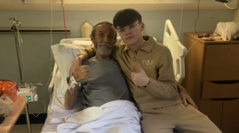 Paul Fairless, left, is in a hospital bed. He is wearing a grey jumper and is smiling. He is laying next to his son, Charlie, who is wearing a beige coloured tracksuit with the words POLO printed in white. Both are showing a thumbs up to the camera.