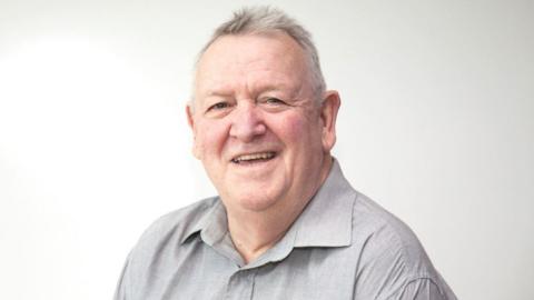 Jeff Lovell wearing a light grey shirt and smiling at the camera. He has short white hair with dark grey patches, and is sitting in front of a white background.