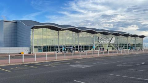The front of Doncaster Sheffield airport. A large glass fronted building with a grey wave-shaped roof.