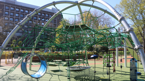 A playground. There is a climbing net in the middle, made of green string. There is a small blue tunnel on the left. There is a rope swing and other playground equipment. Behind the park is a block of flats.