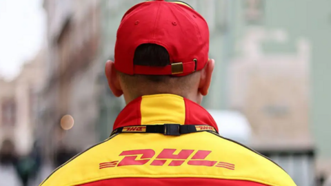 A male DHL worker stands with his back to the camera. His yellow and red uniform has the word DHL on the back and the man is also wearing a red and yellow cap.