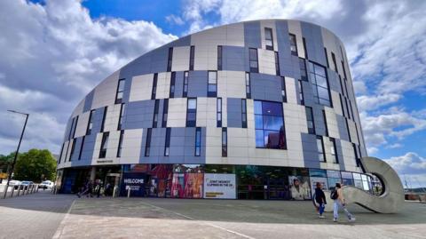The University of Suffolk building. It is a large sloped building covered in windows and blue and white panels. Two people are walking past with others entering one of the doors. A large question mark sculpture is laying down on the right of the image. A blue sky with white clouds is overhead. 