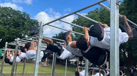 Line of people using parallel bars at an outdoor gym in a park, hanging horizontally and doing pull-up-style exercises, with trees, benches and blue sky visible behind.