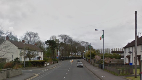 A Google Maps screenshot of Irish Green Street in Limavady.  Terraced houses line parts of both sides of the street and there are trees in the background.  Two cars are driving along the road and there is an Irish tricolour flag on a lamp post. 