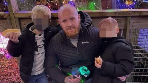 Lenny Scott, who is bald and has a ginger beard, poses with his arms around his young sons (whose faces have been blurred). They are eating hot dogs.