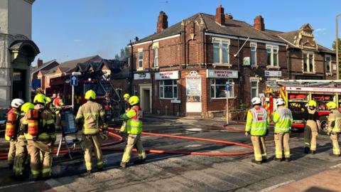 About 10 firefighters wearing light brown uniforms and high-vis jackets and helmets, some with breathing apparatus, stand in a street with two fire engines and red hoses. In the background, smoke can be seen rising into a blue sky from a single-story building with damaged and blackened roof gables. The damaged building is behind a grander, red-brick, two-story building which has signs reading "Swinton WMC".