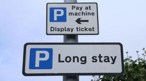 Two signs on a lamppost. The first is a white square sign with a black border. It has a blue square with a white P in. Next to it on the right, it reads "Pay at machine" with a black arrow pointing left. Below, it reads "Display ticket". The second sign which sits below the first is a rectangular white sign with a black boarder. It also has a blue square with a white P. Next to it on the right it reads "Long Stay".