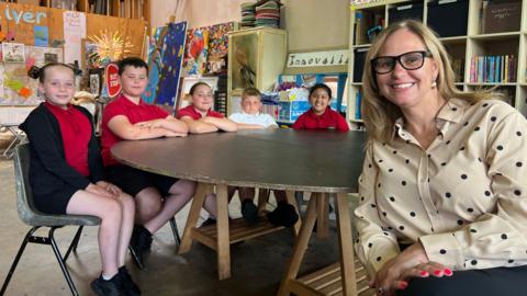 Helen Godwin, the West of England Mayor, is seated smiling with a group of schoolchildren in a brightly lit classroom. The children are gathered around a table, some leaning in with interest. The setting reflects a typical primary school environment, with colourful posters and school supplies in the background.