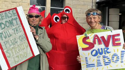 Three campaigners holding placards which say 'please don't pull the plug on Stroud lido' and 'Save our Lido'. The person in the middle is wearing a red lobster costume and the women either side of him are wearing swimming goggles.