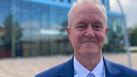 Man smiling at the camera wearing a blue suit jacket, white shirt and blue patterned tie. He has a North Northamptonshire Council lanyard round his neck. In the background is a large glass building.