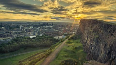 Part of the Radical Road path at the foot of the Salisbury Crags cliffs in Edinburgh's Holyrood Park. The sun is setting over the view of the city centre with Edinburgh Castle visible in the centre of the picture. 