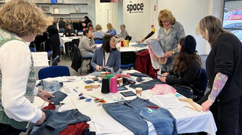 A group of women are using a variation of sewing materials to repair donated jeans. They are standing and sitting round the table talking to each other. 