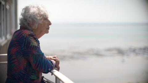 Carol Turansky standing on a balcony. She is wearing a leaf print dress and you can see her side profile. She has grey permed short hair and you can see the sea in the background. She is looking out to the horizon.