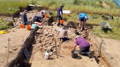 Seven people are bent over digging at an archaeological site. 