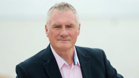 Andrew looks at the camera, wearing a pinstriped pink shirt and a black wool jacket. He has a neat grey moustache and short grey hair.