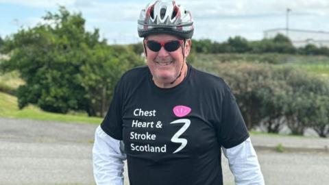 Ken MacAulay is wearing a black t-shirt  over a white cycling shirt. The shirt has the words "Chest, heart and stroke" printed on the chest. He is smiling and wearing a cycling helmet and sunglasses