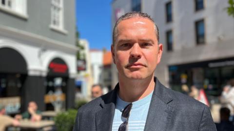 Marcus Calvani wearing a blue jumper and grey blazer. He is standing on a street in town that is blurred out behind him. He is looking into the camera.