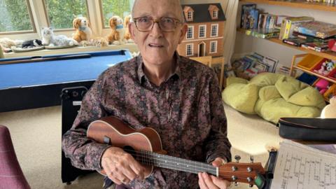 An elderly man sat holding a ukelele wearing a purple patterned shirt and wearing glasses. Behid him a doll's house and books and a bean bag