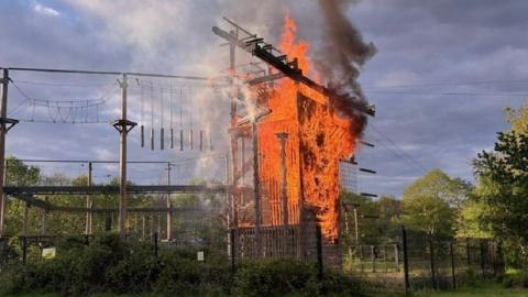 Fencing around a high obstacle course with various structures, wires and netting - a large platform area is engulfed by fire.