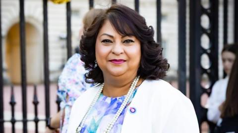 A head-and-shoulders photo of a woman with shoulder-lenght curly brown hair smiling in front of the black gates of Buckingham Palace. She's wearing a flowery dress, a white blazer, and a pearl necklace.