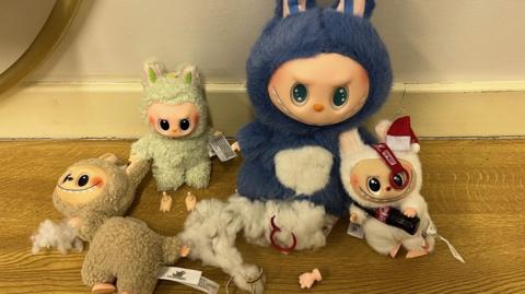 Four furry and colourful fake Labubu dolls on a wooden surface. One of them is broken in half, another has stuffing falling out the bottom. The dolls are small with grinning faces and large eyes. 