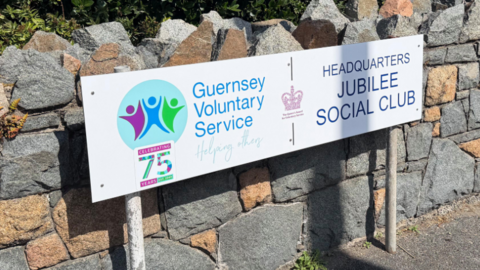 Granite wall with bushes in the background with a white sign displaying the Guernsey voluntary service logo.
