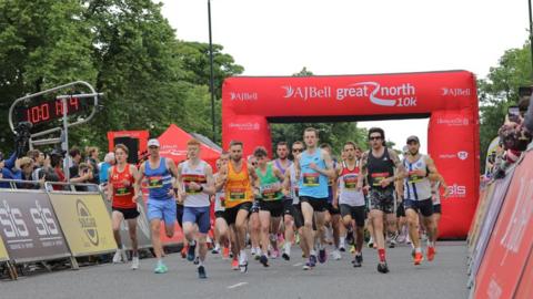 Dozens of runners at the start of a race. The group, mainly men, have passed under a large red starting arch, emblazoned with AJ Bell Great North 10k. Spectators can be seen behind barriers on either side.