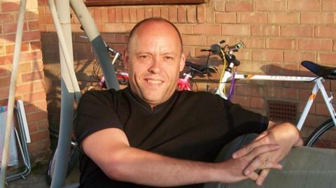 A man in a brown T-shirt sitting in a garden, leaning back on a chair. There are two bicycles leaning against a brick wall behind him.