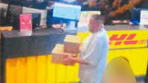 A person stands at a counter holding two cardboard boxes, preparing to ship them. The counter features a bright yellow panel with a red logo, and various signs and equipment are visible on the countertop in the background.