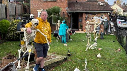 A man in a yellow hoodie and grey shorts standing in a garden with lots of skeletons and other Halloween decorations in it.