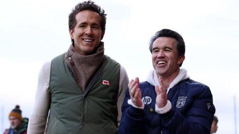 Ryan Reynolds and Rob Mac walk along the touchline at the Cae Ras, with Mac applauding