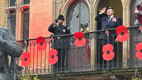 Arthur Lockyear, chairman of Durham City Remembrance Parade, is dressed in a military uniform and stands on a balcony decorated with large poppies at Durham Cathedral. A man also in uniform stands next to him playing the trumpet.